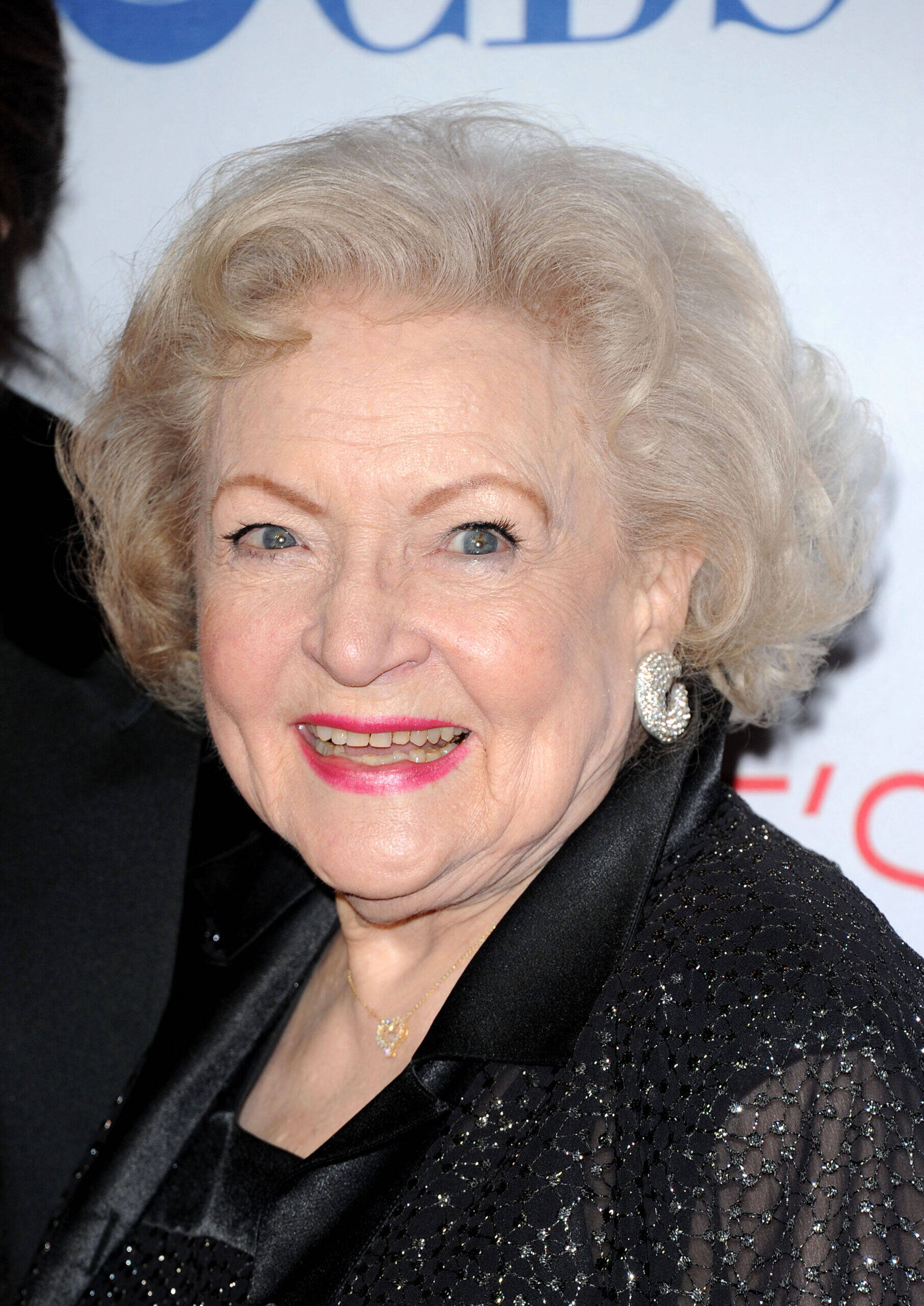 Betty White smiles on the red carpet, wearing a black outfit with sparkling details and silver earrings